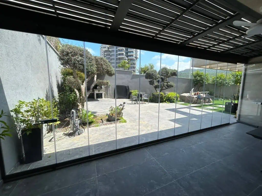 סגירת מרפסת קרקע בזכוכית מתקפלת מחוברת לפרגולה, עם מסילה שטוחה ברצפה ויציאה לגינה מעוצבת